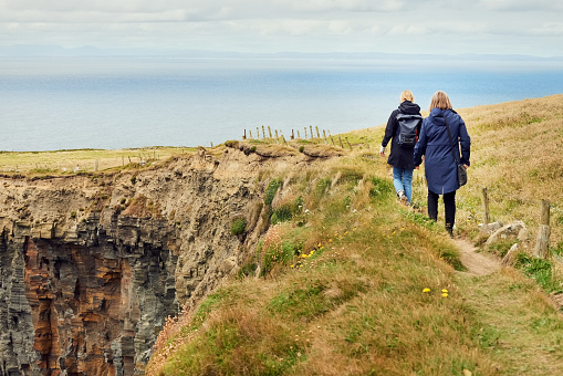 A married couple holding hands while walking along a cliff edge in Polperro, Cornwall against the sea. The woman is looking back at her husband while they walk.