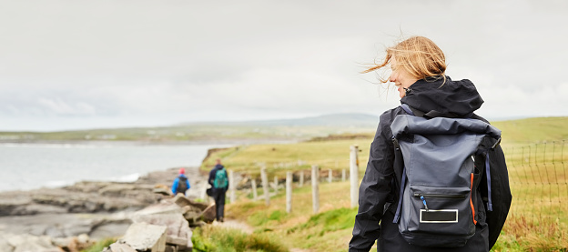 Rear view of a woman walking along a narrow pathway on cliff with windswept hair