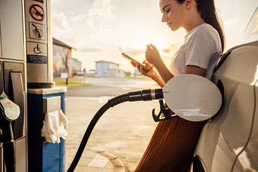 Beautiful young woman text messaging on smart phone while refueling gas tank at fuel pump