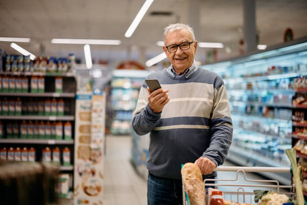 istock Happy mature man using app on mobile phone while shopping in supermarket and looking at camera. 1414488280