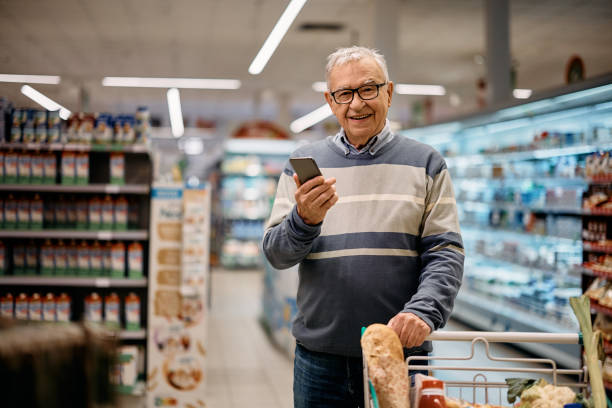 Happy mature man using app on mobile phone while shopping in supermarket and looking at camera. stock photo