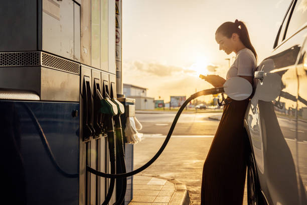 istock Young woman using mobile phone while refueling her car at gas station 1414488385