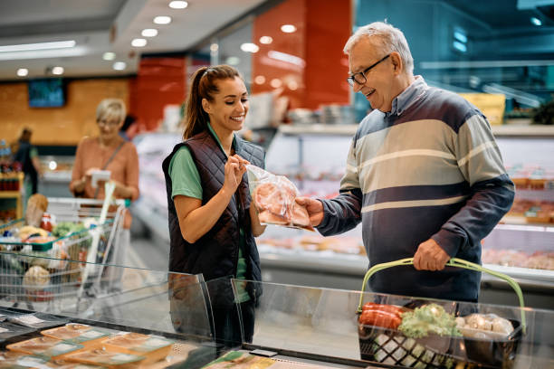 glücklicher supermarktmanager, der den älteren mann bei der auswahl von fleischpaketen unterstützt. - supermarket meat store manager stock-fotos und bilder