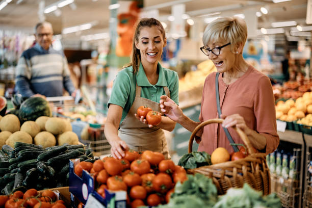 Happy sales clerk assisting senior woman in buying vegetable at supermarket. stock photo
