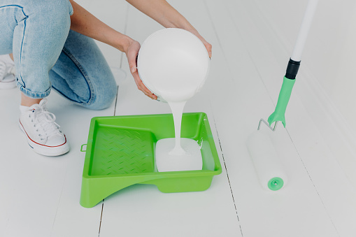 Cropped shot of unrecognizable woman pours white paint in tray, going to paint something with roller, makes house renovation, wears jeans and sneakers. Repair, building, home concept