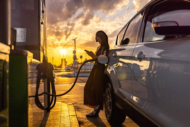istock Young woman using mobile phone while refueling her car at gas station 1414488568