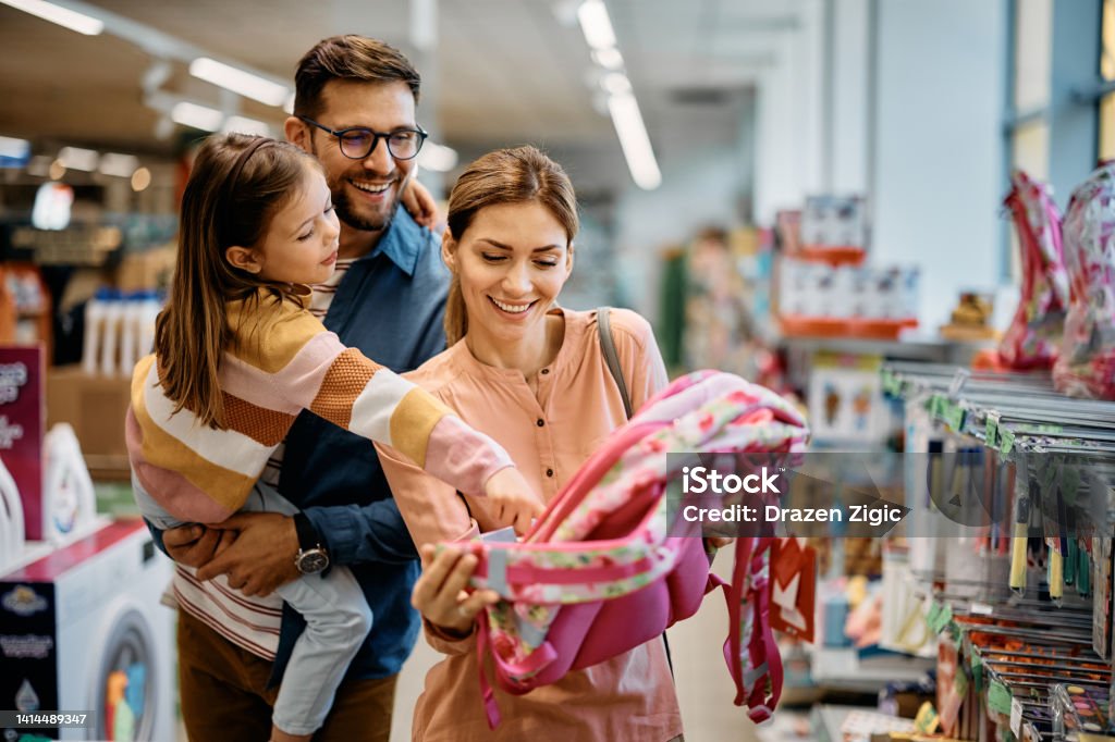 Glückliches kleines Mädchen, das auf den Rucksack zeigt, während sie mit ihren Eltern im Supermarkt Schulmaterial kauft. - Lizenzfrei Einkaufen Stock-Foto Glückliches kleines Mädchen, das auf den Rucksack zeigt, während sie mit ihren Eltern im Supermarkt Schulmaterial kauft. - Lizenzfrei Einkaufen Stock-Foto