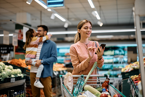 Smiling woman using smart phone while buying groceries in supermarket.