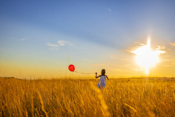 Rear view of girl playing with balloon while running amidst grassy field in meadow against sky during golden hour