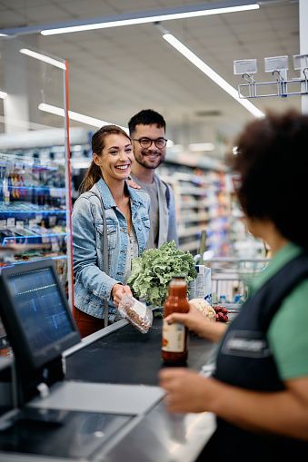 Happy couple buying groceries and talking to supermarket cashier at checkout.