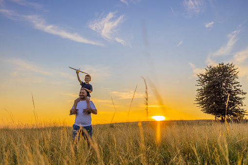 Happy father carrying son with airplane toy while walking amidst grassy field in meadow against sky during sunset