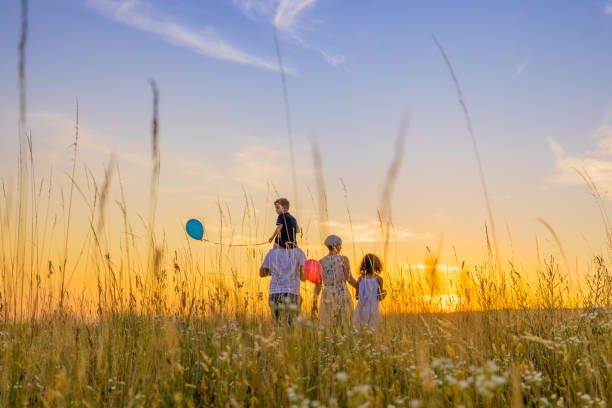 parents et enfants marchant au milieu d’un champ herbeux contre le ciel - sun sky child balloon photos et images de collection