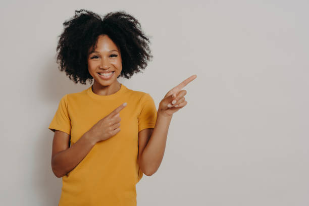 istock Cheerful dark skinned woman dressed in yellow tshirt pointing at left upper corner with forefingers 1414491182