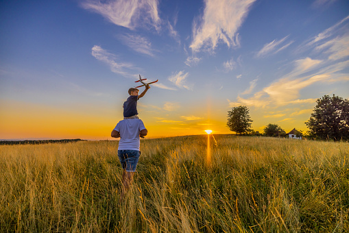 Father carrying son with airplane toy while walking amidst grassy field against sky during sunset