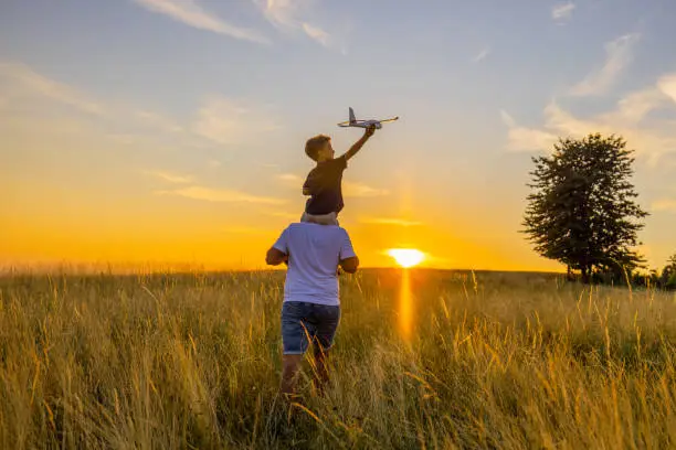 Rear view of father carrying son with airplane toy while walking amidst grassy field in meadow against sky during sunset