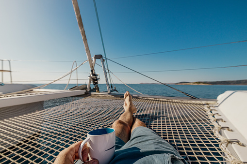 Low section of man having coffee while resting on net at sailboat deck against clear sky during sunny day