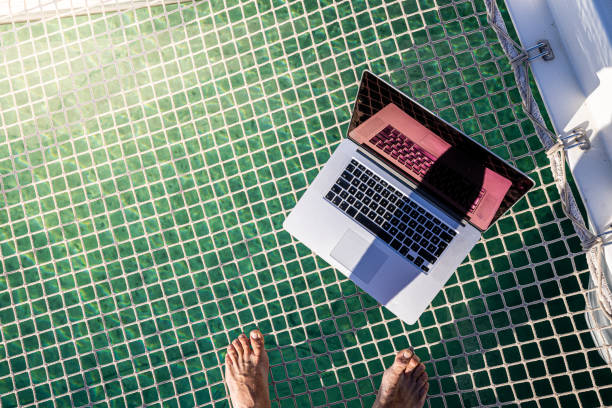On the couch with bare feet resting on the seat, a laptop and hands working on the keyboard. Detail of female hands with an unseen face, white couch, cute and well-groomed bare feet - work from home c