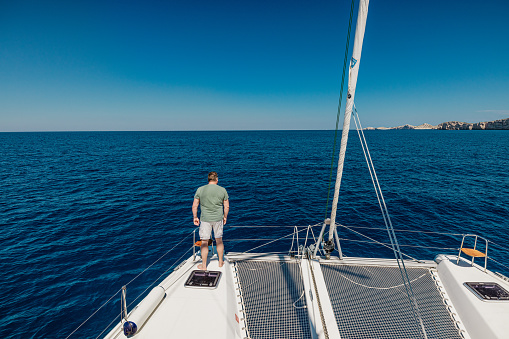 Rear view of man looking at beautiful sea while standing on sailboat against clear blue sky during sunny day