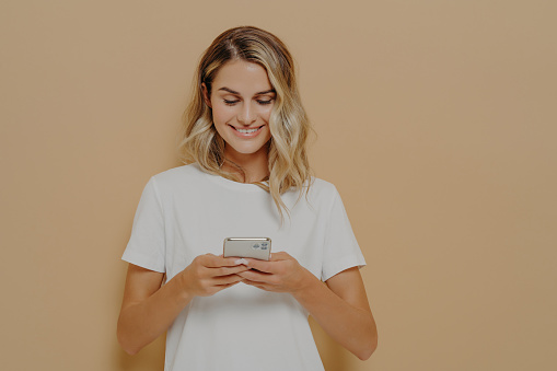 Young happy girl using smartphone, chatting with boyfriend in social media and smiling while standing against orange wall dressed in white t shirt. Modern technologies and networking concept