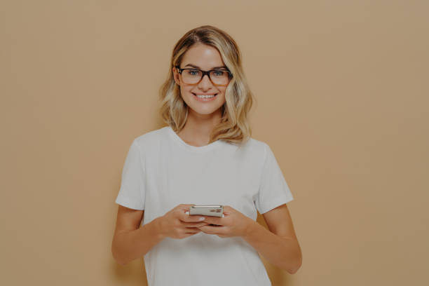 istock Young cute female student in spectacles using cell phone and looking at camera with pleasant smile 1414493359
