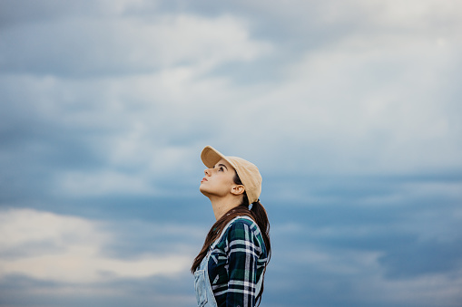 Young female farmer wearing cap looking up while standing against sky