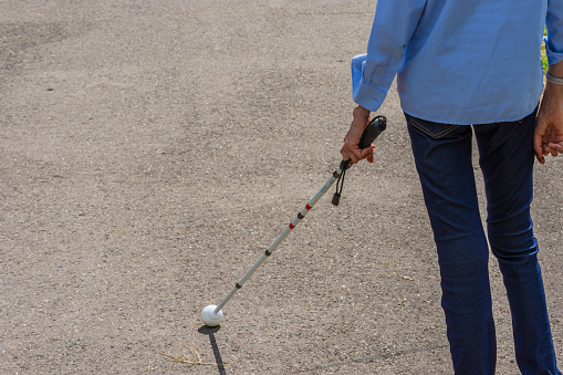 Underfed blind girl with swollen joints and wrists because of arthritis, taking a walk using a white cane on asphalt