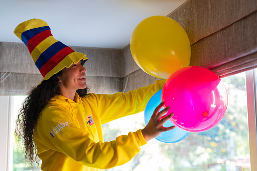 Latin woman with afro hair of average age of 25 years is in the living room of her house dressed in a shirt of the Ecuadorian soccer team and a hat with the flag decorates the house with balloons of team colors