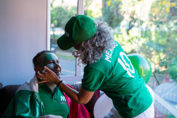 latin couple of older adults are in the living room of their house dressed in a soccer uniform from mexico, a woman makes up the face of her partner with the colors of the team - mexican soccer scores stockfoto's en -beelden