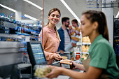 Happy woman putting groceries on checkout counter in supermarket.