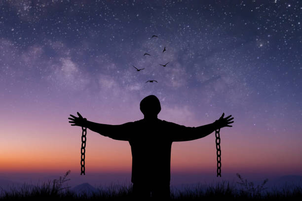 istock Silhouette prisoner stood with his arms outstretched, with a chain broken in his hand standing alone on top of mountain at night time with star, Milky Way and bird over the sky. 1414527427