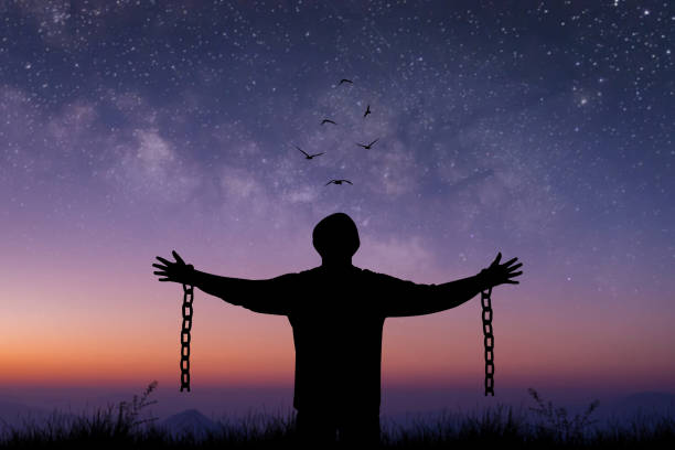 Silhouette prisoner stood with his arms outstretched, with a chain broken in his hand standing alone on top of mountain at night time with star, Milky Way and bird over the sky. stock photo