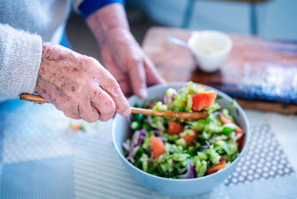 view of elderly woman's hands stirring the salad with a wooden spoon on the table of the kitchen stock photo