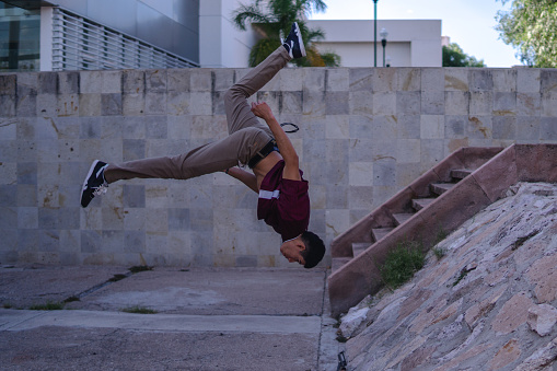 Latino man dressed in urban clothing doing a somersault in the air. He takes in the air of a young Hispanic man doing parkour. Latin boy in a park doing gymnastics. Hispanic man in an acrobatic pose.