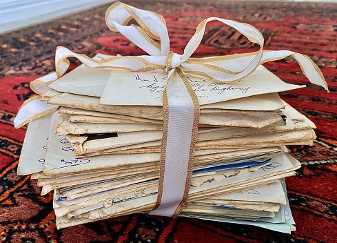 Horizontal still life of 56 year old stack of Australian posted hand written love letters from married couple tied with white and gold ribbon sitting on red Persian rug