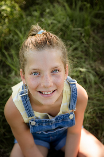 A young girl standing, leaning on a tree wearing dungarees and a long sleeved t-shirt. She is holding a magnifying glass while looking at the camera and smiling.