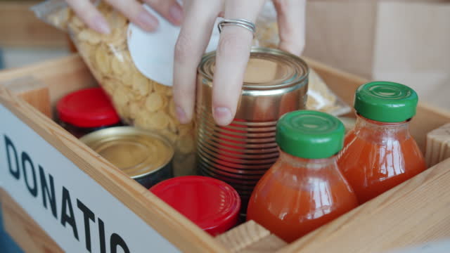 Close-up of wooden box with food inside and human hands volunteers putting provision for charity