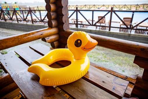 A yellow inflatable duck, lies on massive wooden table in cozy gazebo of one of houses at recreation center against the backdrop of a large pool covered with heavy pouring rain