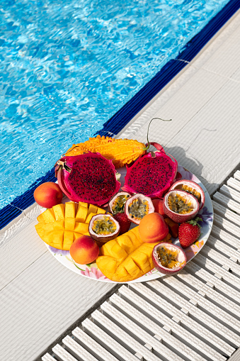 Plate with tropical fruits near the pool. Mango, dragon fruit, strawberry, passion fruit, apricot, momordika on a plate on a blue pool background. Thai fruits.