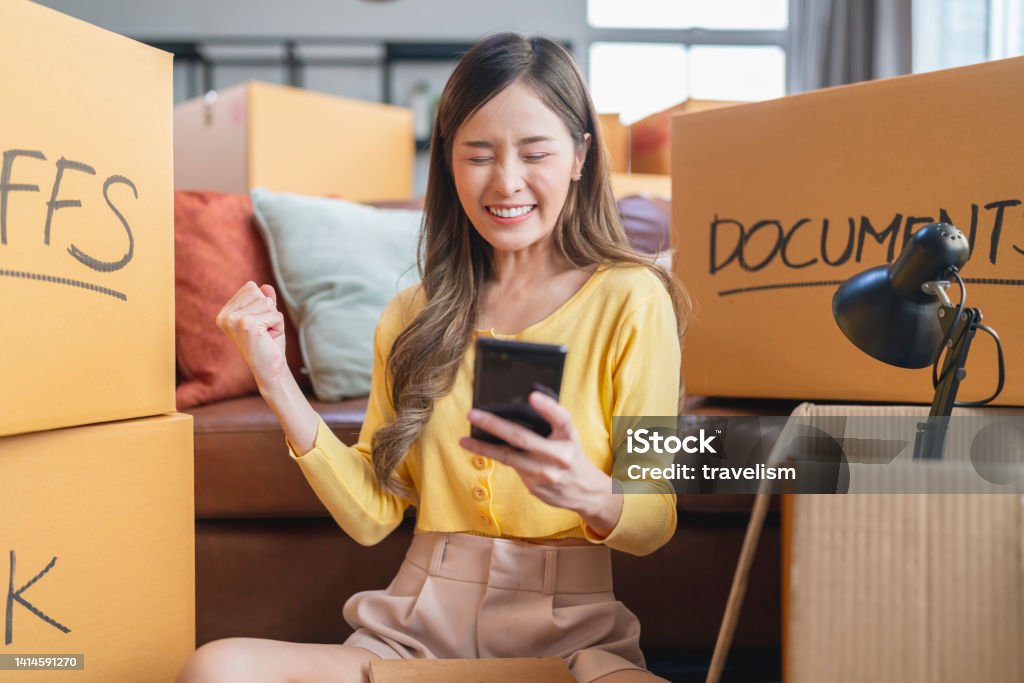 asian mature adult female woman is packing and unpacking as she is moving into a new place house. she is using her smartphone to talk and chat text to friends and family or finding a delivery service nearby.home moving ideas concept Decluttering Stock Photo