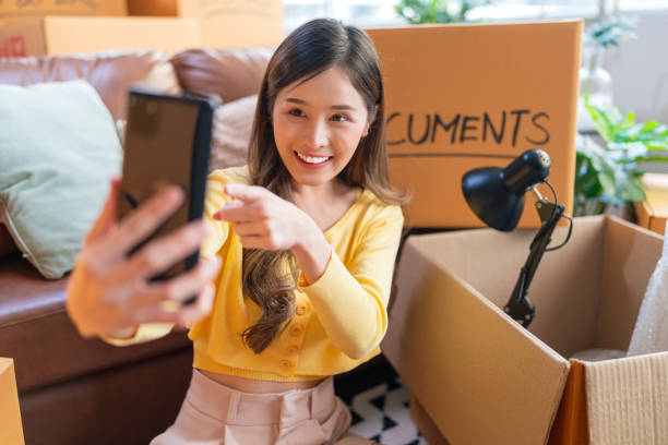 asian mature adult female woman is packing and unpacking as she is moving into a new place house. she is using her smartphone to talk and chat text to friends and family or finding a delivery service nearby.home moving ideas concept la mujer adulta madura asiática está empacando y desempacando mientras se muda a una nueva casa de lugar. ella está usando su teléfono inteligente para hablar y chatear mensajes de texto con amigos y familiares o encontrar un servicio de entrega cerca.
