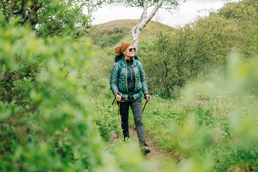 Full length of a mature woman walking through on mountain trail. Female in hiking gear walking in nature and looking at the scenery.