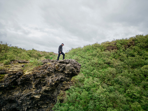 View of a mature man standing on the cliff edge and looking down with green mountain in background. Mature man looking at the view from edge of a mountain.