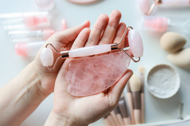 Woman holding gua sha tool and cosmetic over white table, top view