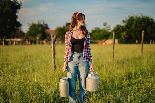 Beautiful young female farmer carrying milk canisters while standing in green grassy rural field