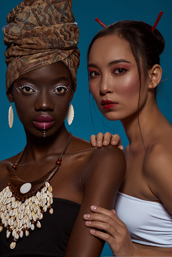 Cropped of attractive multiethnic girl friends in their national traditional styles looking at camera. Black girl wearing african outfit and accessories with asian woman. Blue background. Studio shoot