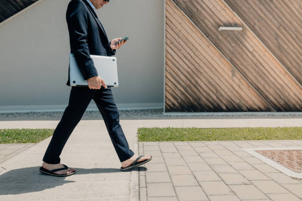 istock Businessman using mobile phone while walking with laptop on footpath by wall 1414602527