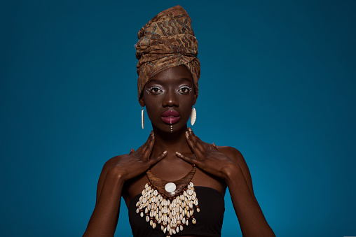 Front view of stylish black girl looking at camera. Attractive young woman wear tank top and traditional african turban, necklace and earrings. Female beauty. Blue background. Studio shoot. Copy space