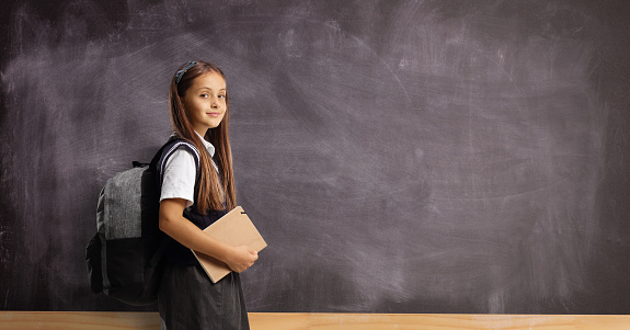 Schoolgirl with a backpack and book standing in front of a blank blackboard and looking at camera