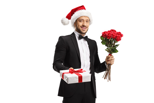 Handsome man in a suit and bow tie wearing a santa claus hat and holding roses and gift box isolated on white background