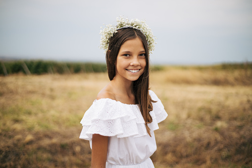 Smiling little girl in angel image with gold wings posing in greenery forest, happy looking away. Funny young lady in white dress in sunshine woodland. Fairytale, fantasy concept. Copy ad text space